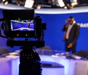 Broadcast camera focused on a presenter in a modern studio. The camera screen shows the presenter standing behind a desk, while the studio backdrop and lighting are visible in soft focus.